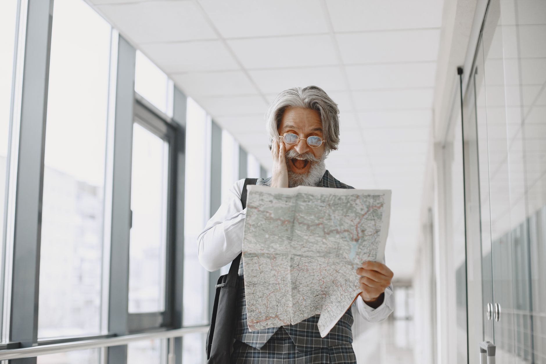 Elderly man with gray hair and eyeglasses looking surprised while holding a map indoors.
